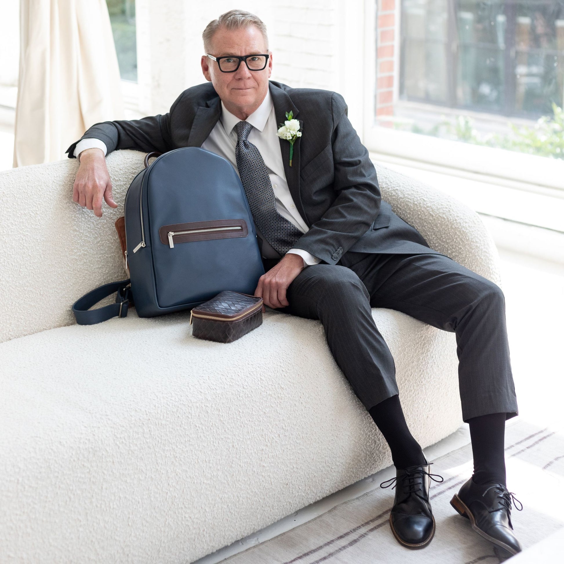A man is sitting on a white couch next to a Moto Large backpack in Textured Peacock.