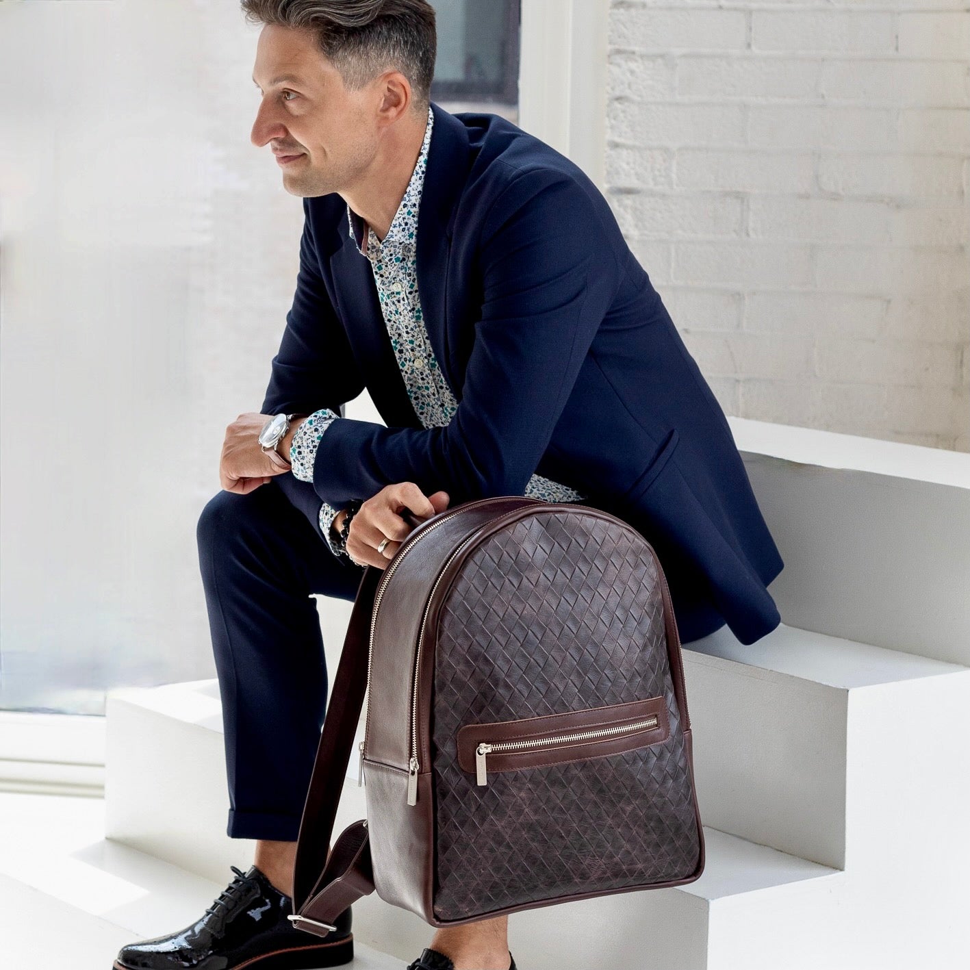 A man i sitting down on a staircase holding a Moto Large backpack in Basketweave Brown/Espresso.