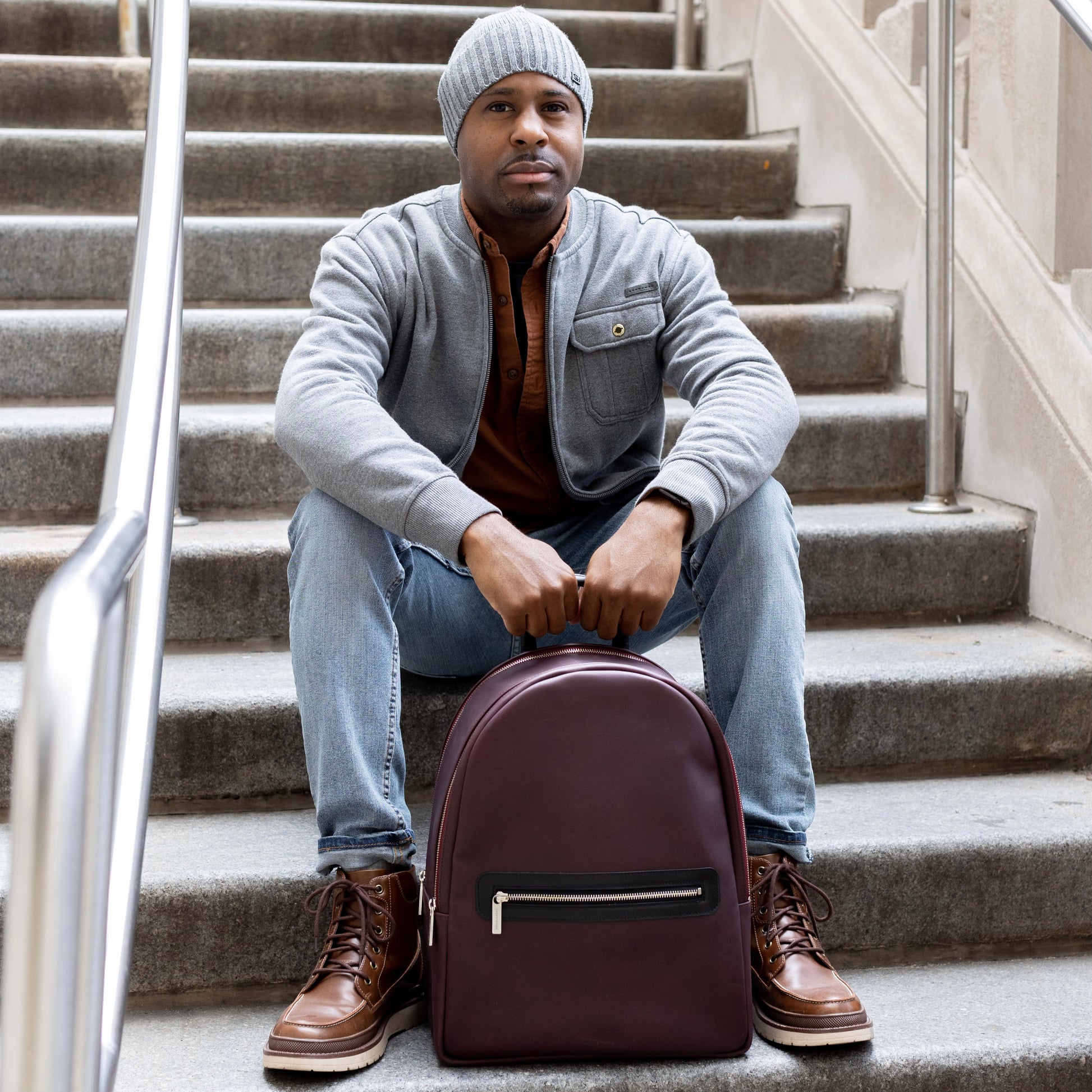 A man is sitting on a staircase outside, holding a Moto Large backpack in the color Oxblood.